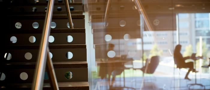 a metal staircase on the left forms into a glass wall reflecting building angles and orange light with two people working at tables in the background