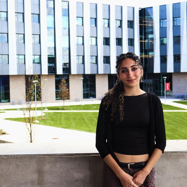 a young femal student with long brown hair wearing a black shirt and pants stands in front of a ledge with a large gray building in the background