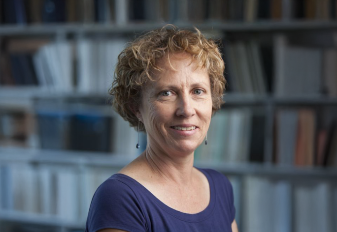 A color photo of woman with short curly hair. Books in background.
