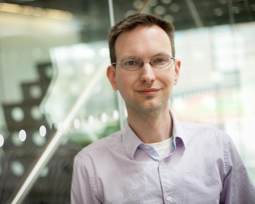 man with short, brown hair and glasses, leans against glass wall